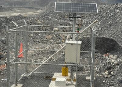 Pequeña instalación cercada con panel solar sobre un poste, caja eléctrica y cono naranja en un entorno rocoso tipo cantera.