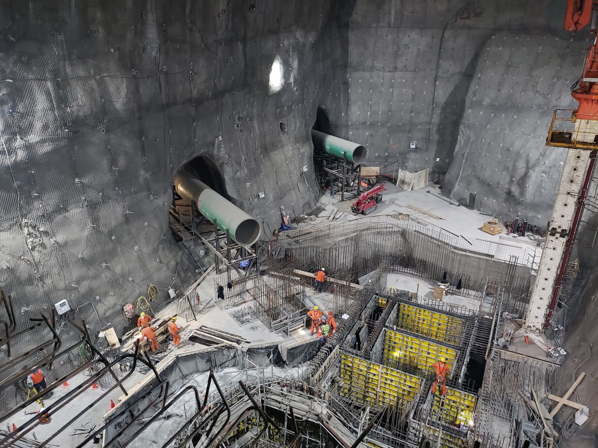 Fotos Sectores-1_1 Obrero y estructura de obra subterránea con túneles en pared rocosa, grandes tubos verdes y encofrados metálicos con trabajadores con chalecos naranjos.