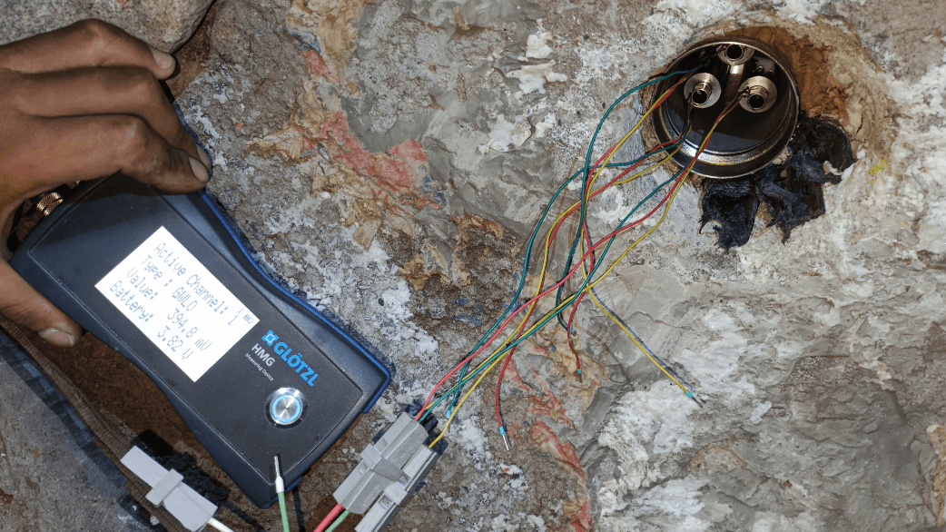 extensometros_2 Mano sosteniendo medidor Glötzl HMG con pantalla que muestra valores eléctricos, cables multicolores conectados a dos conectores grises sobre superficie rocosa.