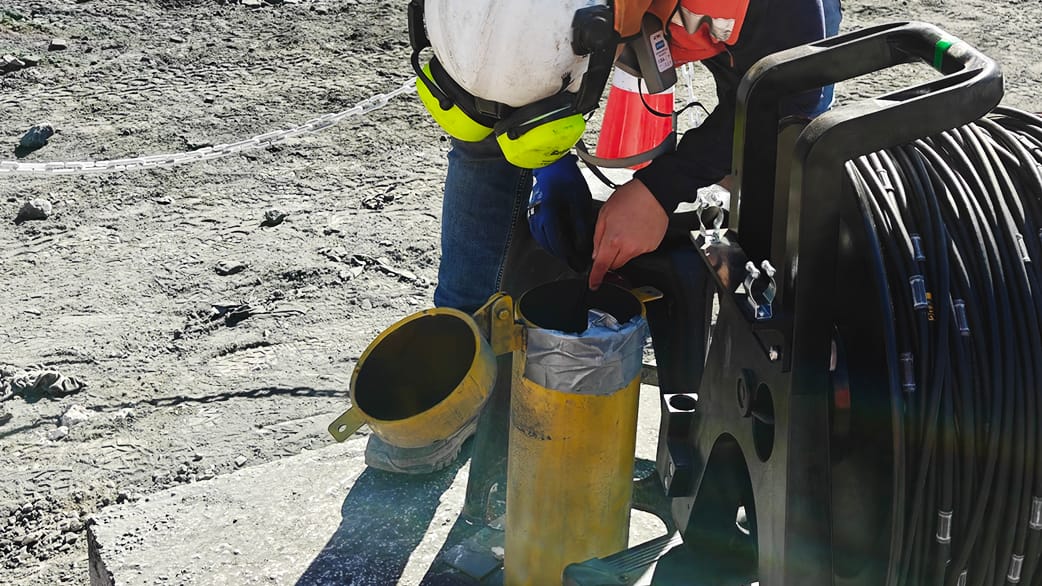 oplus_2097184 Trabajador con casco blanco, chaleco naranja y protección auditiva revisando equipo de perforación junto a un carrete grande de cable en obra al aire libre