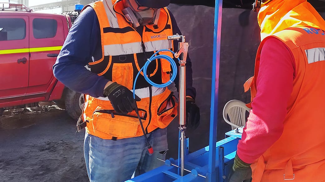 inclinometros_4 Dos trabajadores con chalecos y cascos de seguridad naranjas manipulando equipo y cables azules en una zona de obra.